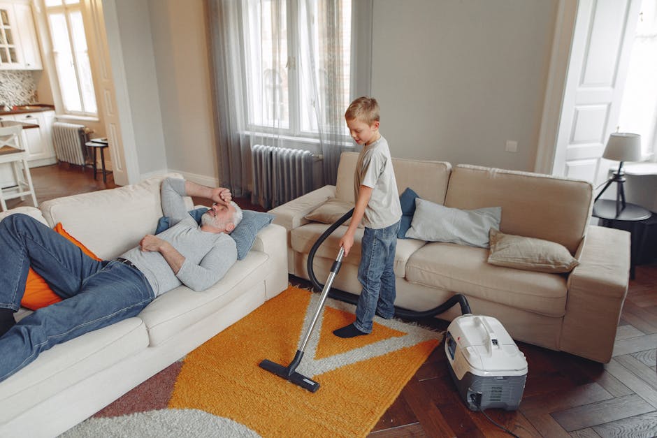 A professional cleaner dressed in white protective coveralls, a hairnet, face mask, and blue shoe covers, performing deep carpet cleaning in a bright, modern living room. The person is using a upright