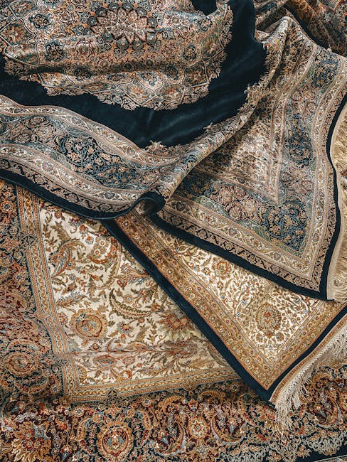 Close-up image of intricately patterned Persian-style rugs featuring detailed floral and geometric motifs in shades of beige, gold, navy blue, and brown, layered on a hard floor surface. The rugs are